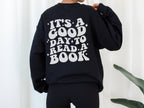 woman wearing black sweatshirt with back print that says it's a good day to read a book standing in front of white curtain