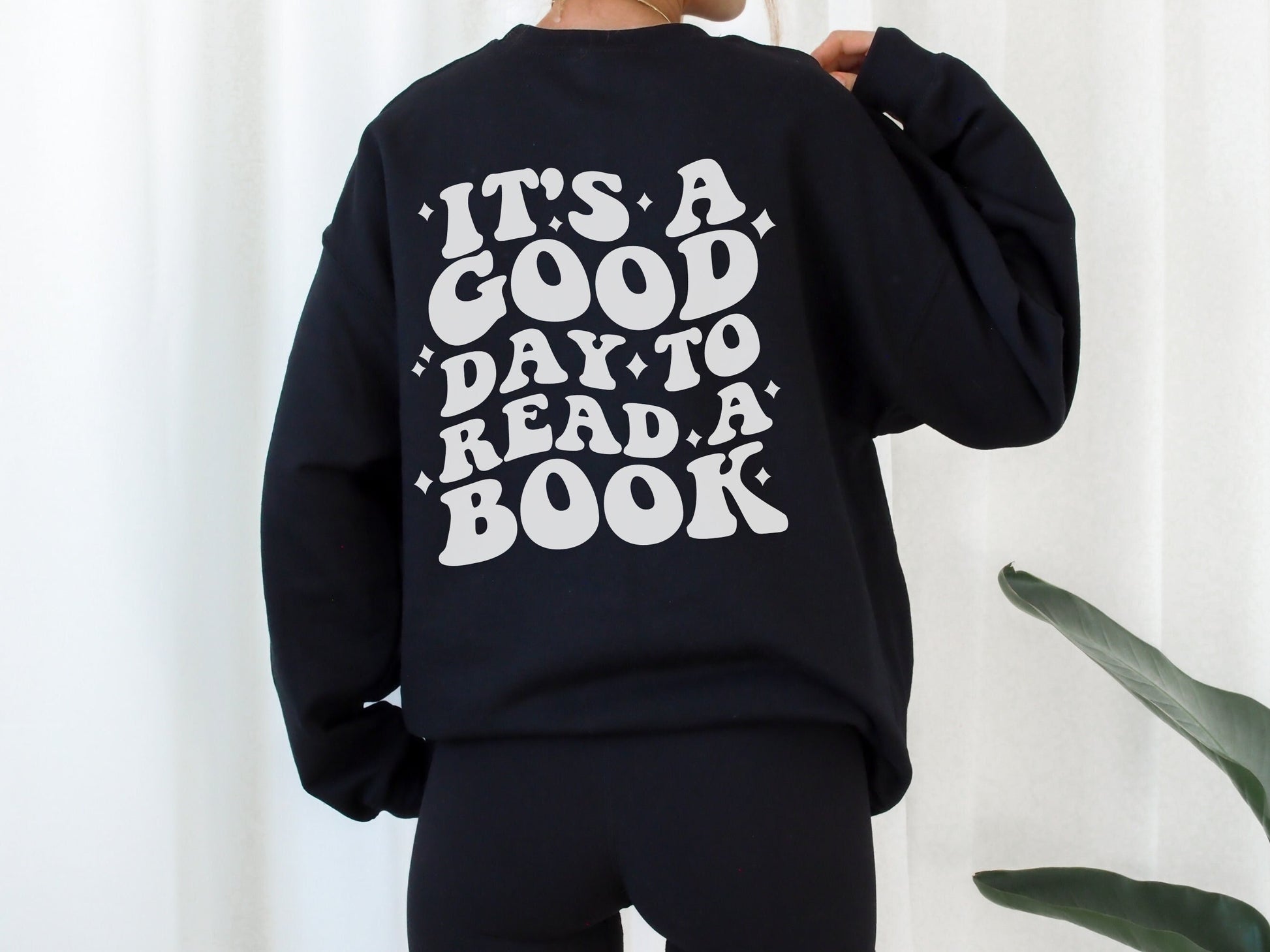 woman wearing black sweatshirt with back print that says it's a good day to read a book standing in front of white curtain