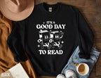 black sweatshirt with it's a good day to read text and two cartoon book characters, styled with jeans, hat, and coffee