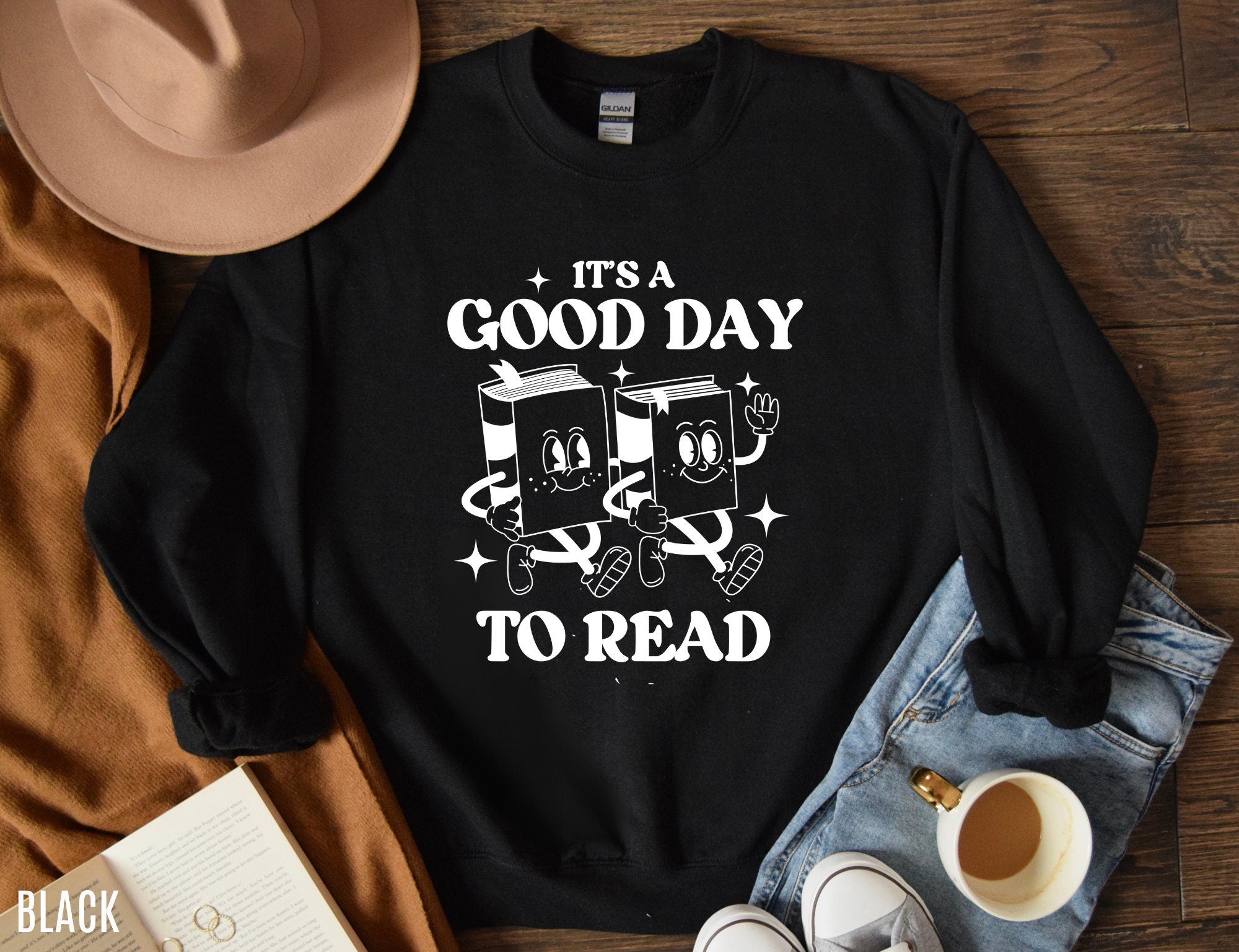 black sweatshirt with it's a good day to read text and two cartoon book characters, styled with jeans, hat, and coffee