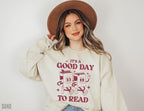 woman wearing sand-colored sweatshirt with it's a good day to read graphic and tan hat, smiling at camera