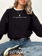woman wearing black sweatshirt that says it's a good day to write sitting cross-legged in jeans and sneakers