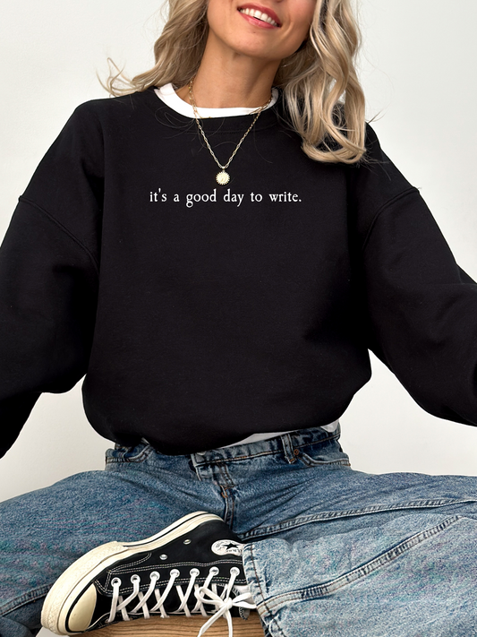 woman wearing black sweatshirt that says it's a good day to write sitting cross-legged in jeans and sneakers