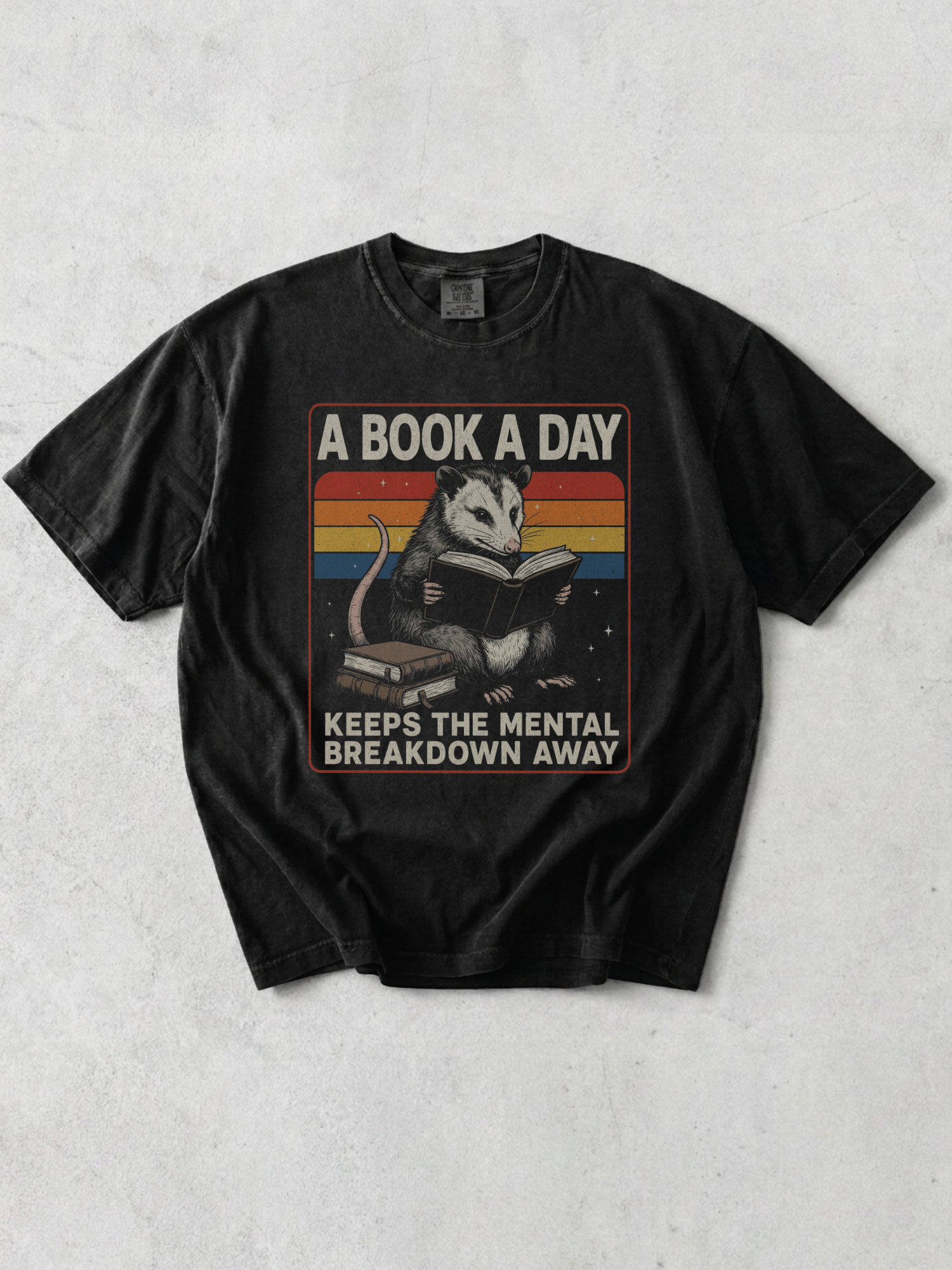 Flat lay of graphite Comfort Colors t shirt with opossum reading a book and retro rainbow design with text a book a day keeps the mental breakdown away on neutral background