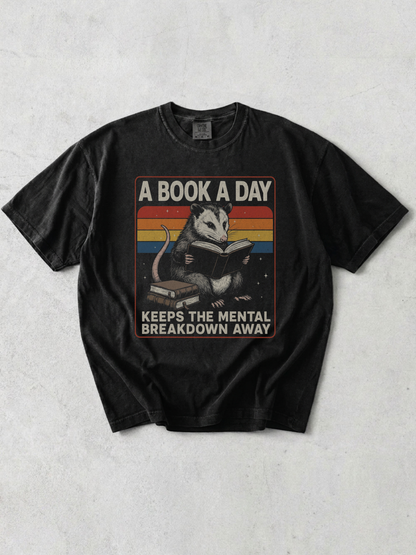 Flat lay of graphite Comfort Colors t shirt with opossum reading a book and retro rainbow design with text a book a day keeps the mental breakdown away on neutral background