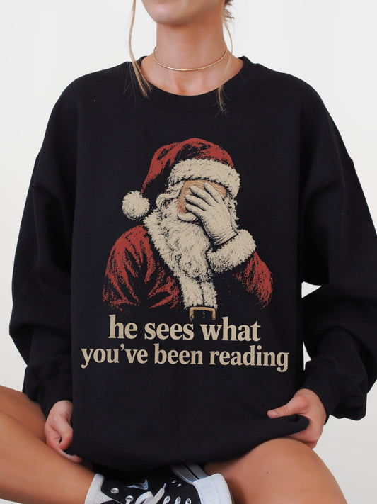 Black Gildan sweatshirt featuring vintage Santa covering his eyes with the funny bookish phrase he sees what you’ve been reading, a Christmas sweatshirt for readers and book lovers