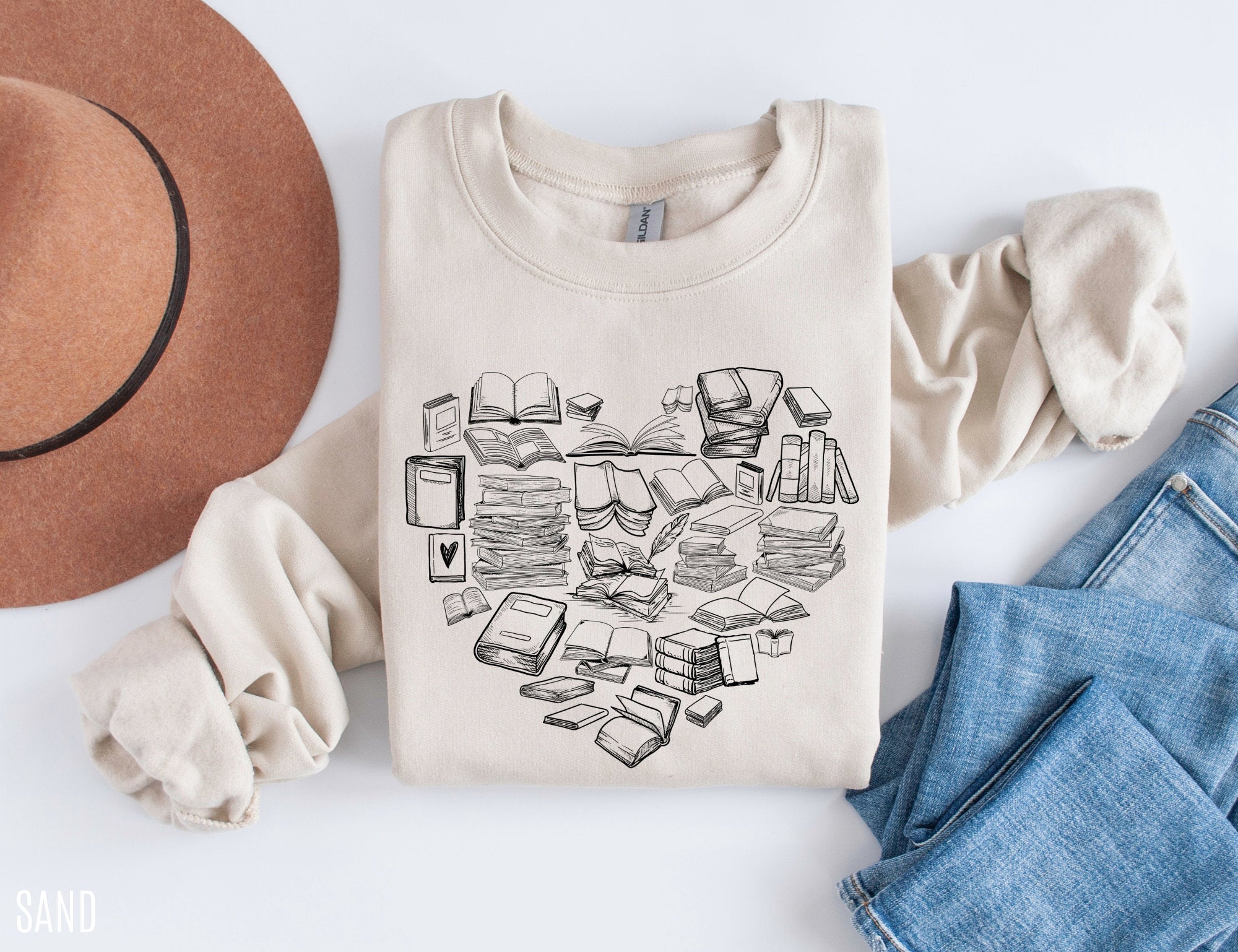 folded sand-colored sweatshirt with hand-drawn book stacks in a heart shape, styled with hat and jeans
