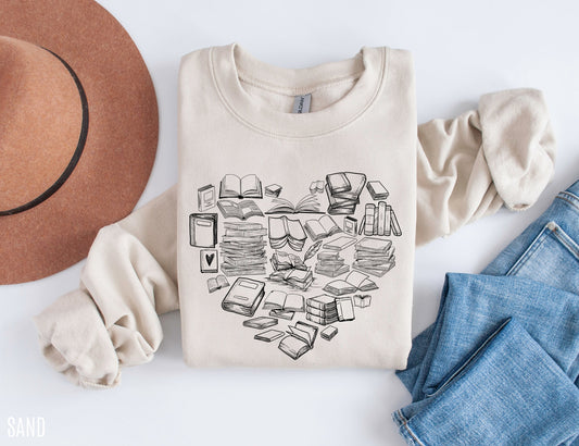 folded sand-colored sweatshirt with hand-drawn book stacks in a heart shape, styled with hat and jeans