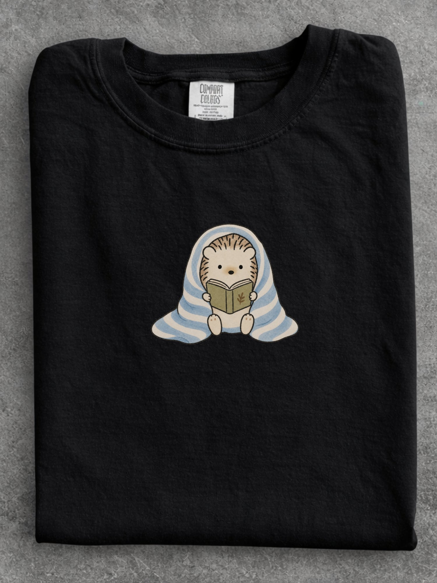 Black Comfort Colors shirt featuring a cute hedgehog reading a book while wrapped in a blue striped blanket, cozy bookish shirt for readers, book lovers and bookworms