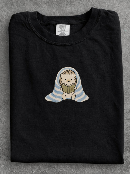Black Comfort Colors shirt featuring a cute hedgehog reading a book while wrapped in a blue striped blanket, cozy bookish shirt for readers, book lovers and bookworms