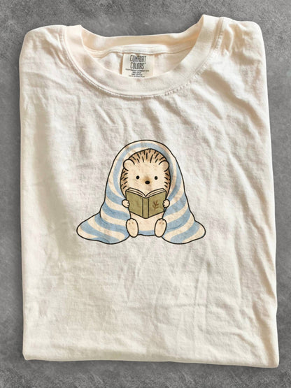 Ivory Comfort Colors shirt featuring a cute hedgehog reading a book while wrapped in a blue striped blanket, cozy reading graphic tee and bookworm gift for book lovers