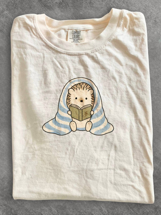 Ivory Comfort Colors shirt featuring a cute hedgehog reading a book while wrapped in a blue striped blanket, cozy reading graphic tee and bookworm gift for book lovers