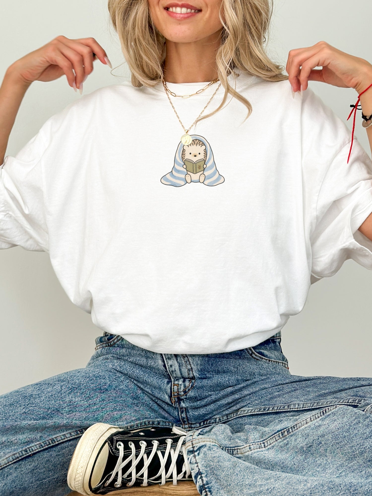 White Comfort Colors shirt featuring a small cute hedgehog reading a book while wrapped in a blue striped blanket, minimalist bookish shirt for readers and book lovers
