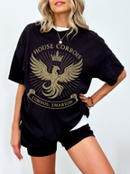 Model wearing a black House Corbois graphic tee with a bold golden phoenix crest and crown design. Styled with layered jewelry and black biker shorts, this shirt is perfect for fantasy book lovers who rep their favorite house.