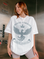 Model wearing a white House Corbois t-shirt featuring a large vintage-style phoenix crest with crown and rays, inspired by the realm of Luminos, Emarion. The shirt is styled casually with biker shorts for a comfy fantasy look.