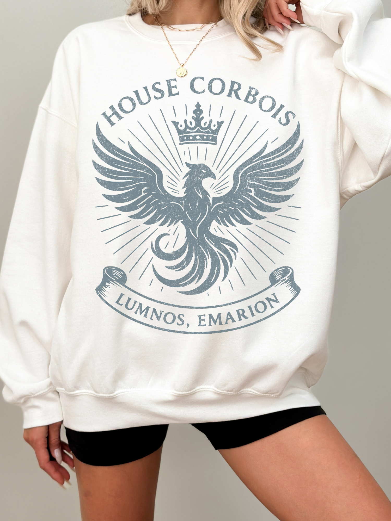 Woman in ivory sweatshirt with large faded blue phoenix crest, reading “House Corbois – Lumnos, Emarion”

