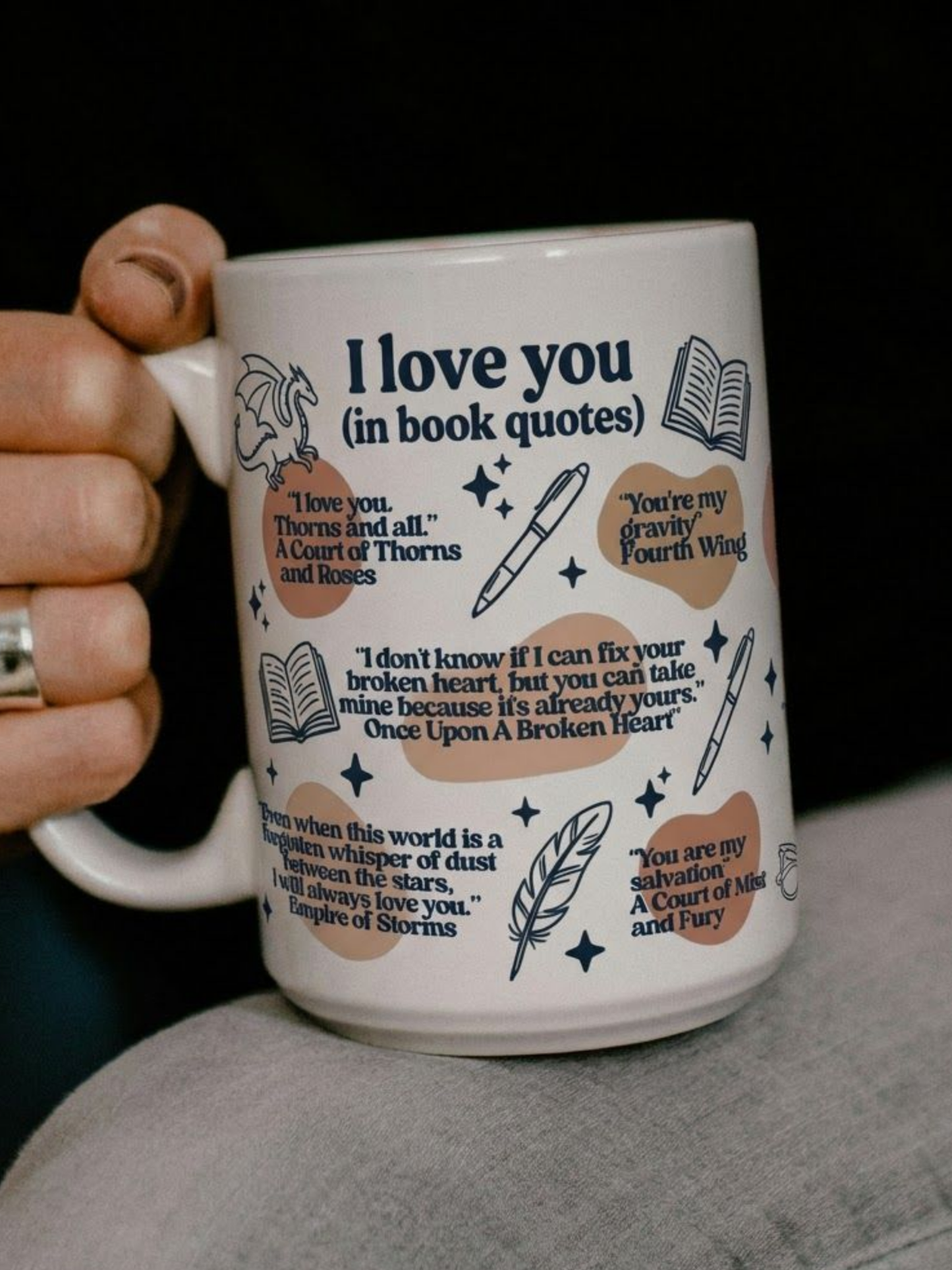 White ceramic I Love You in Book Quotes mug held in hand featuring romantic fantasy book quotes with dragon, book, feather, and star illustrations on cozy dark background