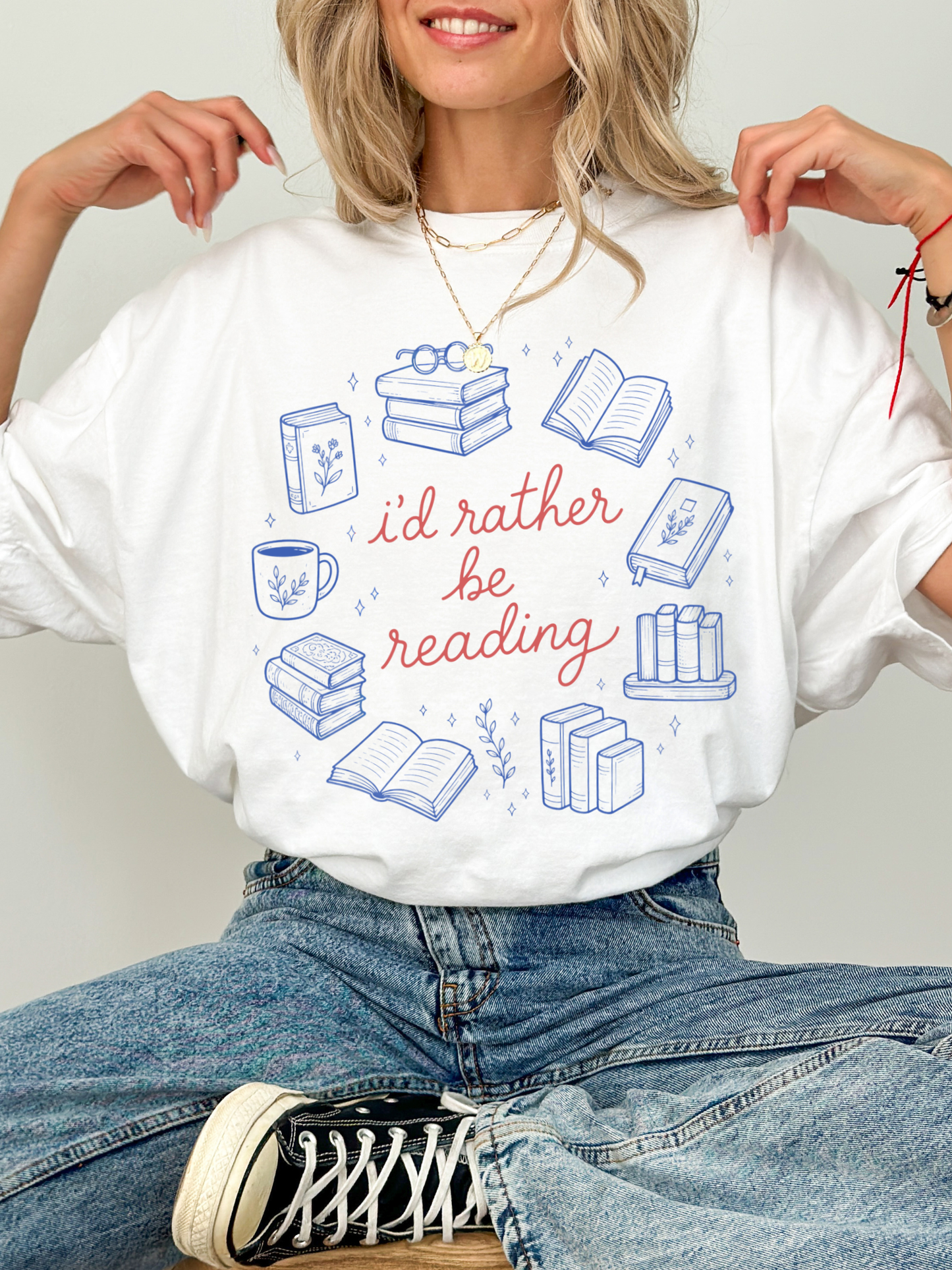 woman sitting in jeans wearing oversized white bookworm graphic t shirt with id rather be reading design and blue illustrated book graphics