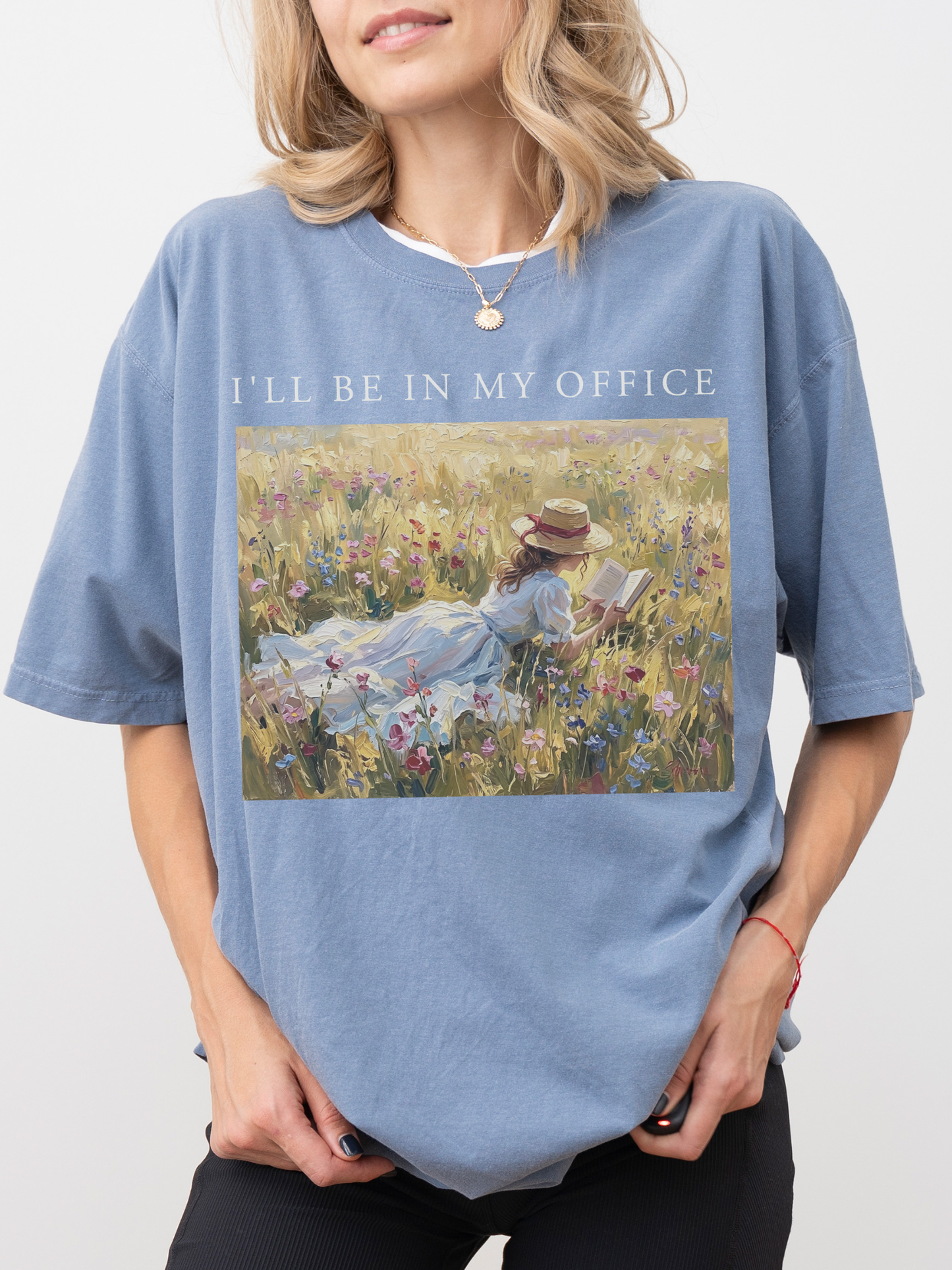 Woman wearing a faded blue t shirt with I’ll be in my office text and a soft impressionist style artwork of a girl reading in a sunny flower field