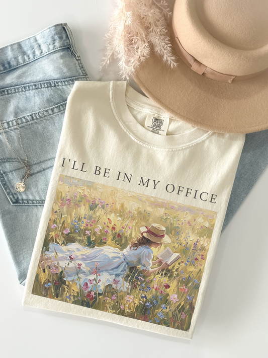 Flat lay of a ivory t shirt with I’ll be in my office text and a vintage meadow reading scene styled with denim jeans and a beige hat