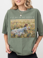 Woman wearing an oversized moss green t shirt with I’ll be in my office text and a soft vintage painting of a girl reading in a floral meadow