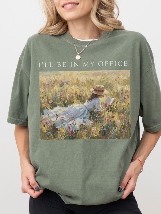 Woman wearing an oversized moss green t shirt with I’ll be in my office text and a soft vintage painting of a girl reading in a floral meadow