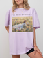 Woman wearing a orchid oversized t shirt with I’ll be in my office text and a vintage style painting of a girl reading in a wildflower meadow