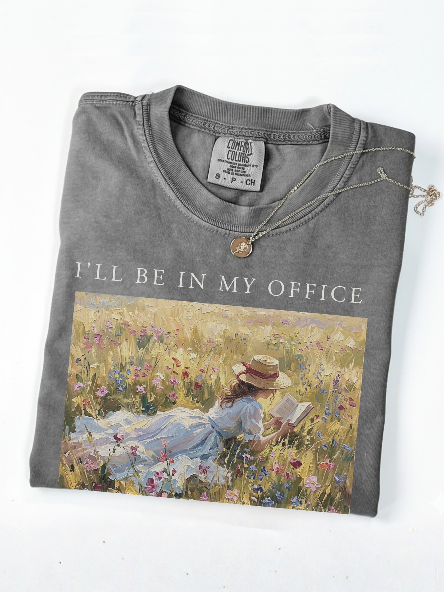 Folded pepper t shirt with I’ll be in my office text and an impressionist painting of a girl reading a book while lying in a field of wildflowers