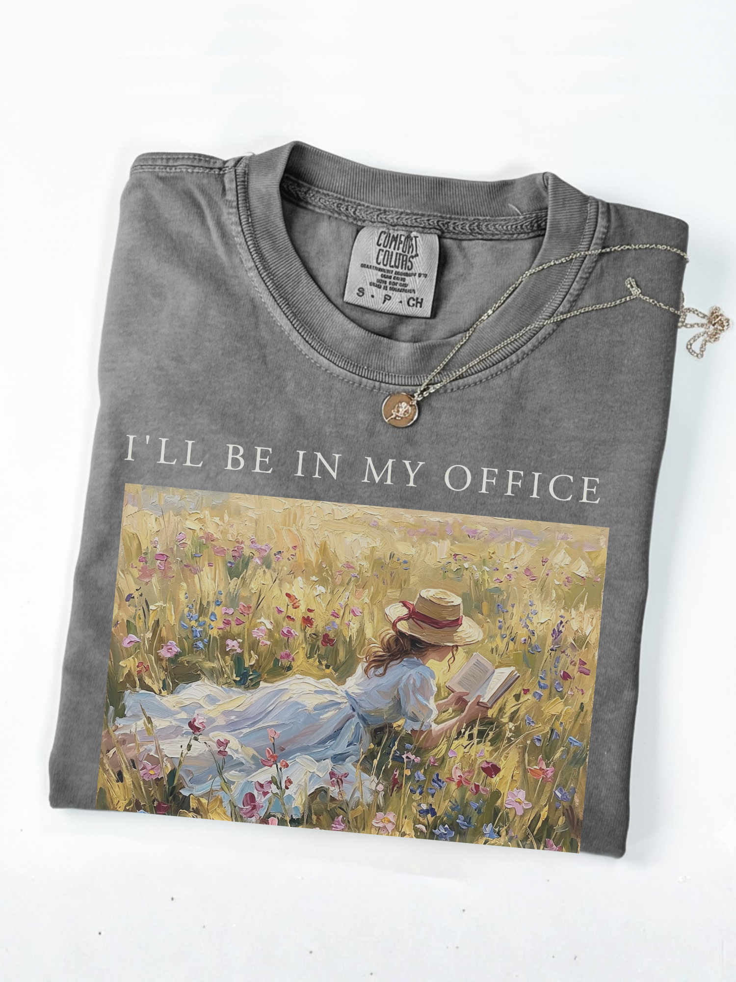 Folded pepper t shirt with I’ll be in my office text and an impressionist painting of a girl reading a book while lying in a field of wildflowers