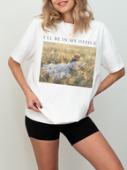 Woman wearing a white t shirt with I’ll be in my office text and a romantic impressionist artwork of a girl reading in a sunlit wildflower field