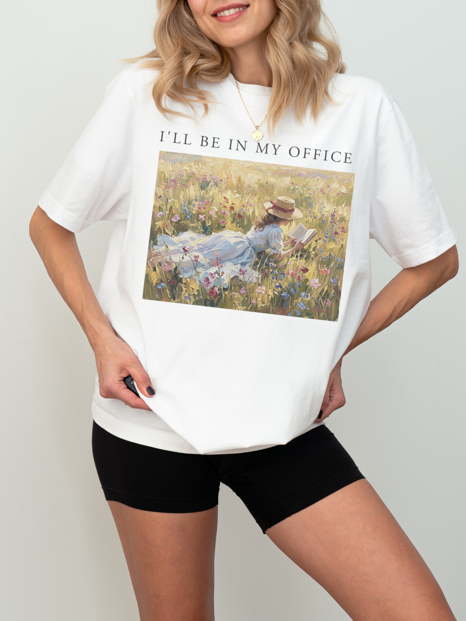Woman wearing a white t shirt with I’ll be in my office text and a romantic impressionist artwork of a girl reading in a sunlit wildflower field