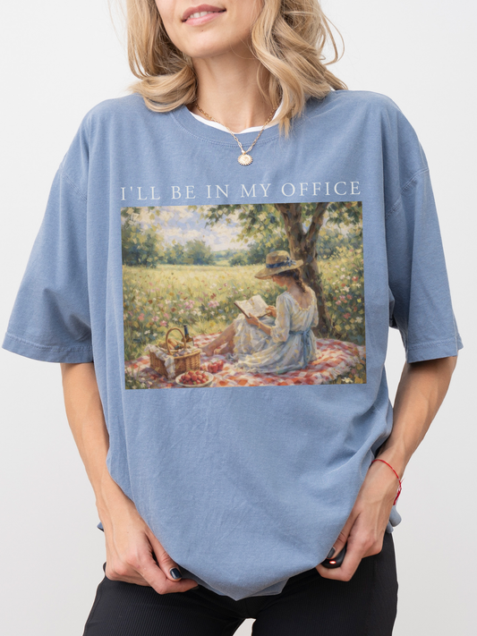 Woman wearing a faded blue jean t shirt with I’ll be in my office text and a soft vintage style illustration of a woman reading outdoors in a field with flowers
