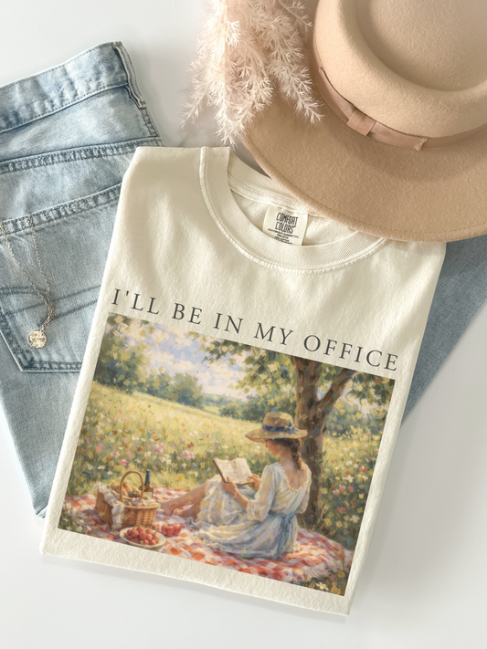 Flat lay of a ivory t shirt with I’ll be in my office text and a vintage picnic reading scene styled with denim jeans and a beige hat