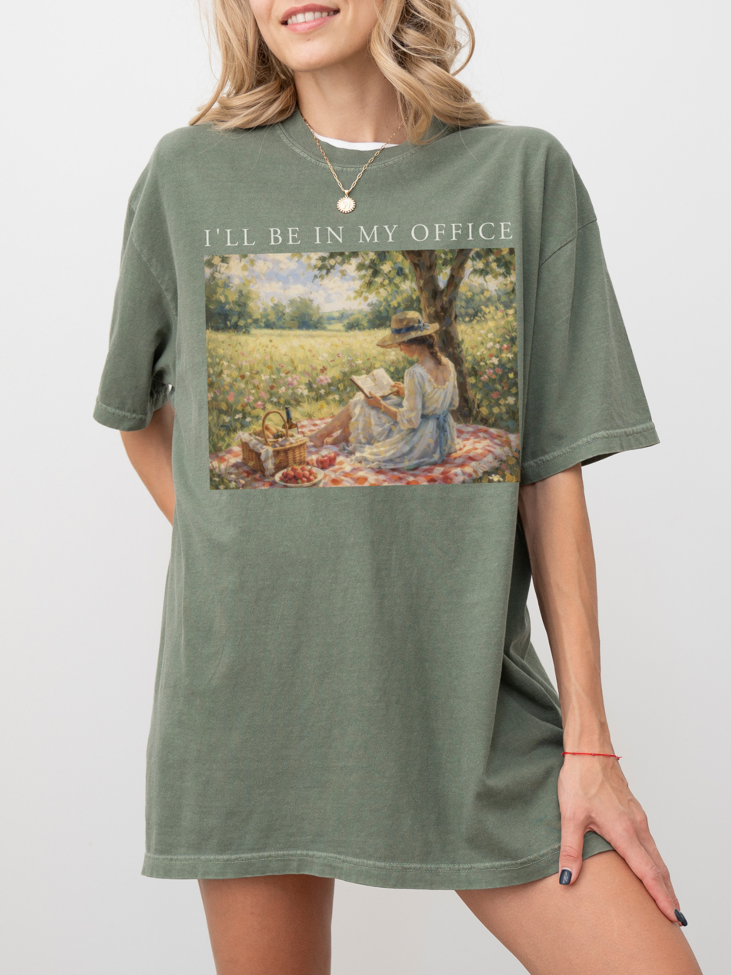 Woman wearing an oversized moss green t shirt with I’ll be in my office text and a vintage style illustration of a woman reading on a picnic blanket outdoors