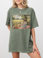 Woman wearing an oversized moss green t shirt with I’ll be in my office text and a vintage style illustration of a woman reading on a picnic blanket outdoors