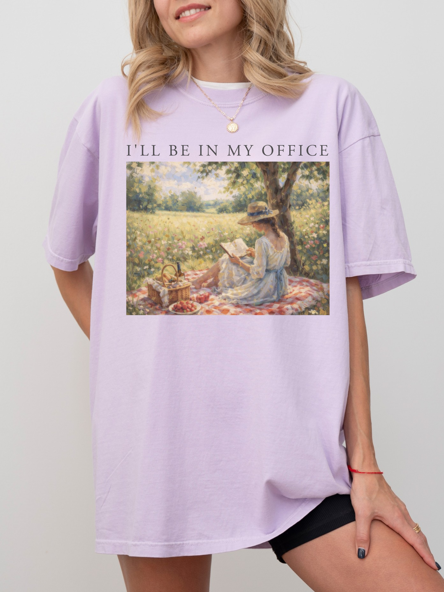 Woman wearing a orchid oversized t shirt with I’ll be in my office text and a vintage painting of a woman reading a book on a picnic blanket in a sunny meadow