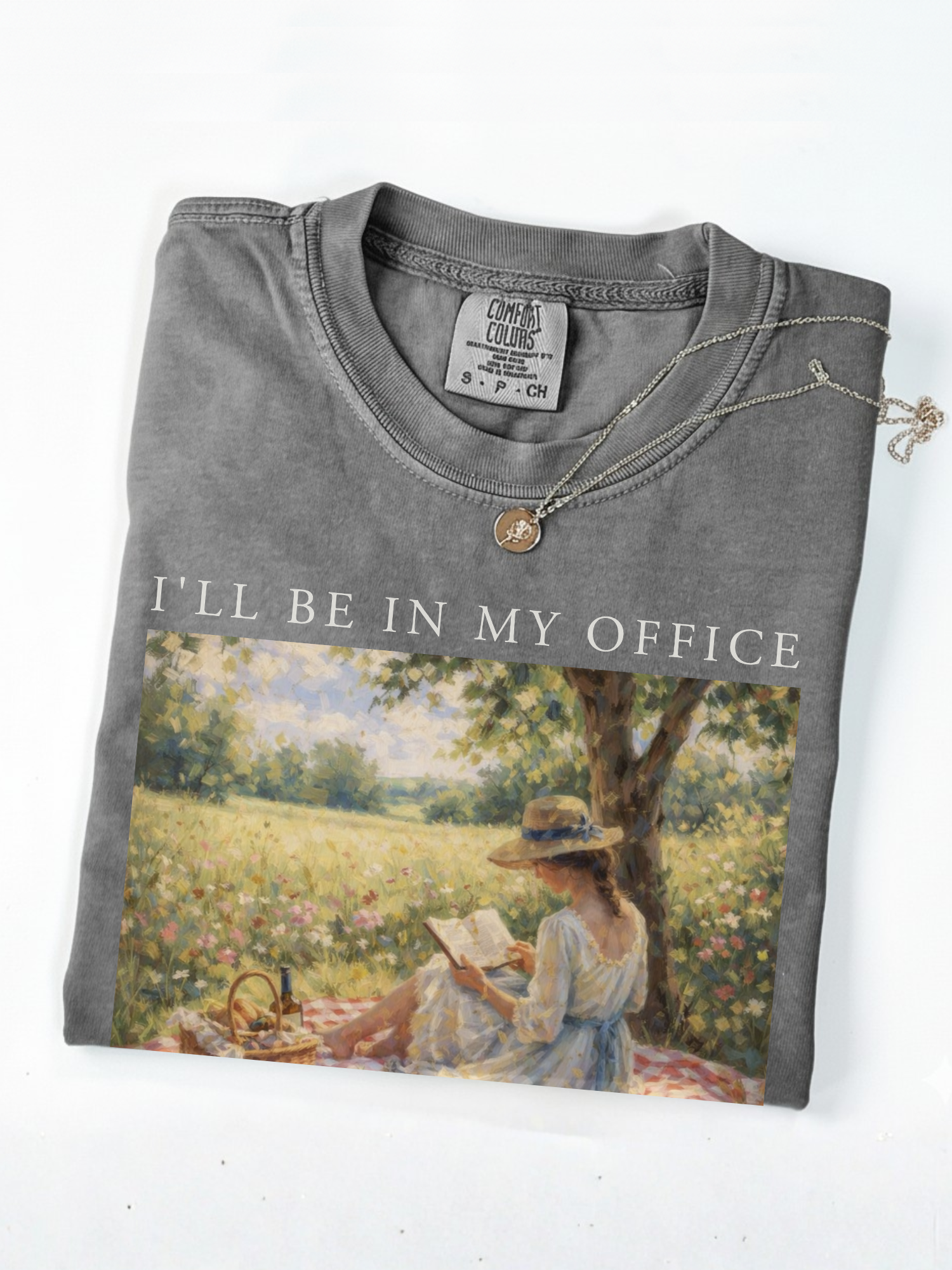 Folded pepper t shirt with I’ll be in my office text and a soft vintage painting of a woman reading under a tree in a floral meadow