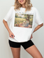 Woman wearing a white t shirt with I’ll be in my office text and a romantic vintage painting of a woman reading a book in a sunlit meadow picnic setting