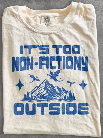 A folded cream-colored cotton t-shirt with a blue graphic print that reads 'IT'S TOO NON-FICTIONARY OUTSIDE' above and below a blue mountain, pine trees, and dragon illustration. The inner neck label shows the brand 'Comfort Colors'.