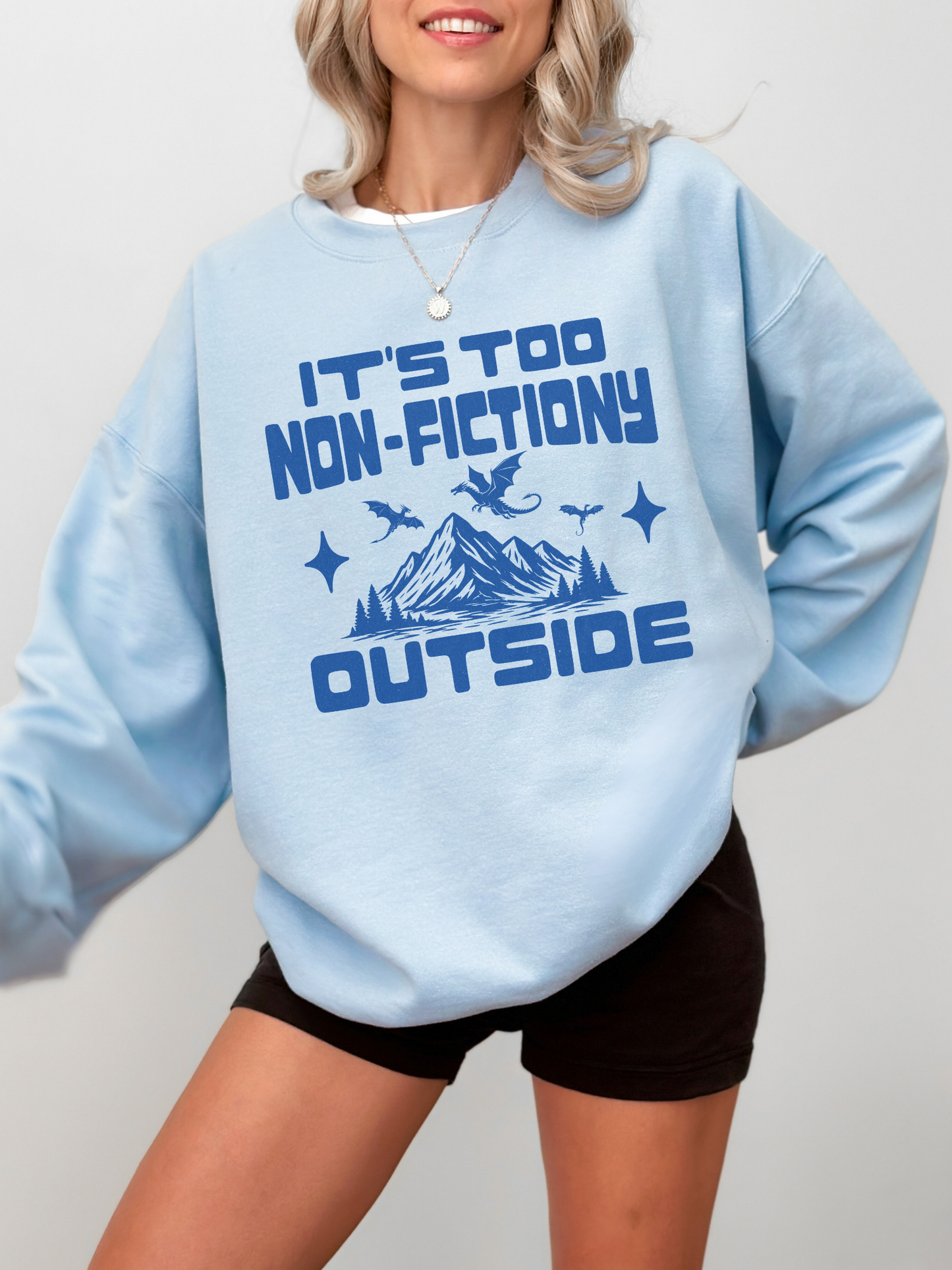 A woman wearing a light blue crewneck sweatshirt with a dark blue graphic that reads 'IT'S TOO NON-FICTIONARY OUTSIDE' above a mountain and dragon illustration, paired with black shorts against a light gray background.