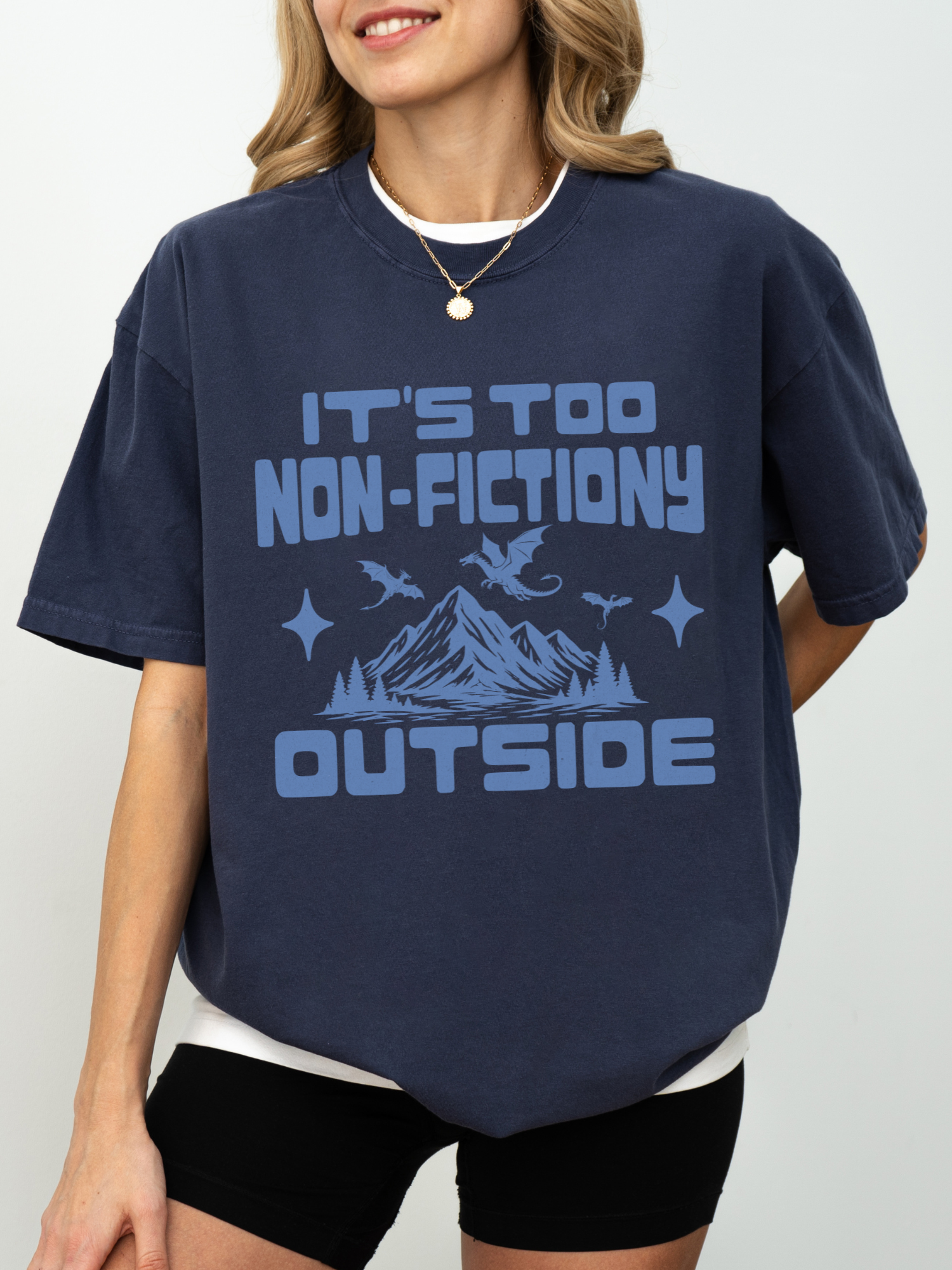 A woman wearing an oversized navy blue graphic t-shirt with a light blue print that reads 'IT'S TOO NON-FICTIONARY OUTSIDE' with a mountain and dragon illustration, paired with black bike shorts against a light gray background.