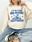 A woman wearing a cream-colored sweatshirt with a blue graphic that reads 'IT'S TOO NON-FICTIONY OUTSIDE' with a mountain and bird design, paired with blue jeans and black sneakers.