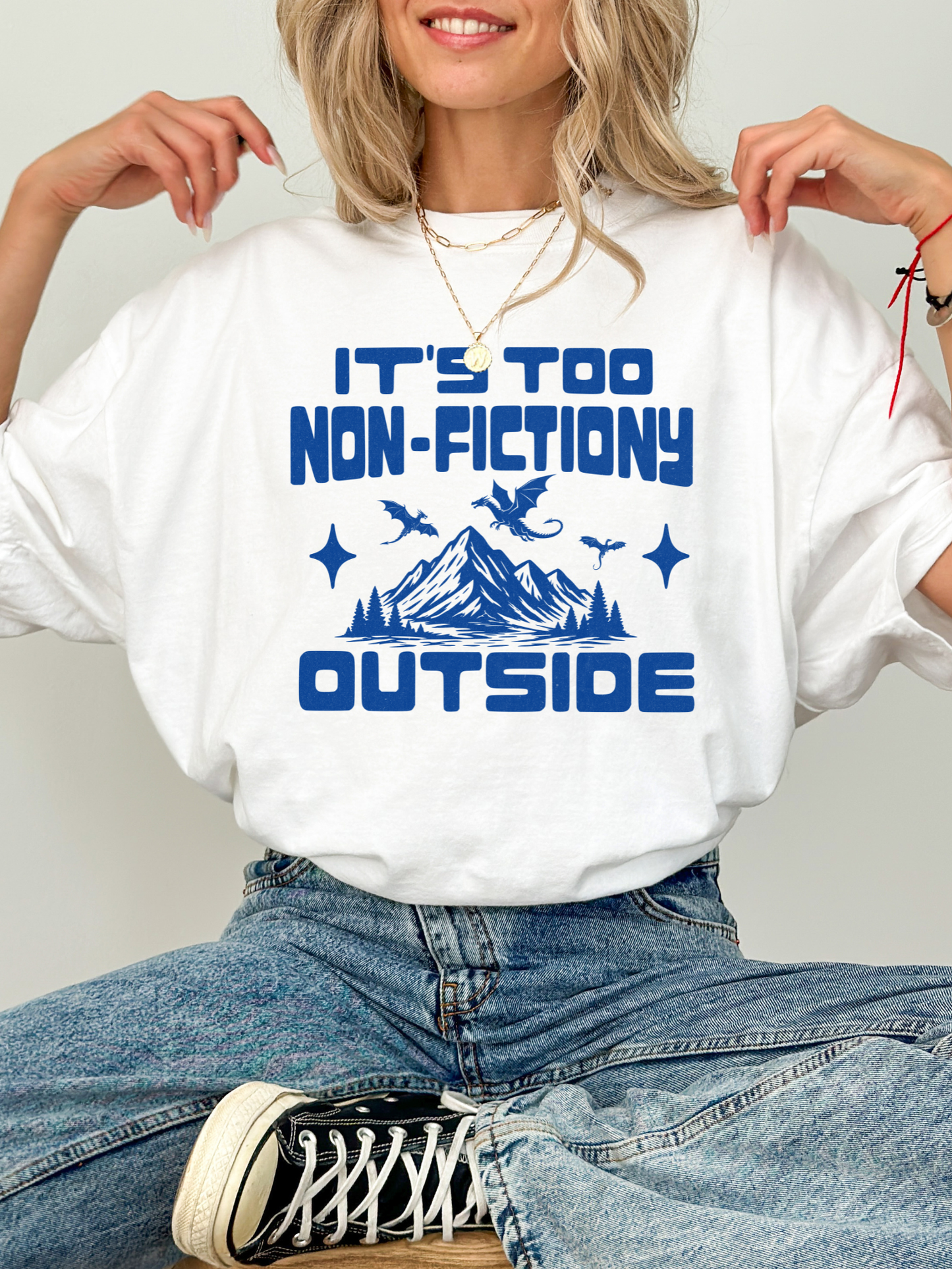 A woman wearing a white graphic t-shirt with blue text reading 'IT'S TOO NON-FICTIONARY OUTSIDE' and a blue mountain/creature illustration, paired with blue jeans and a black-and-white sneaker against a light gray background.