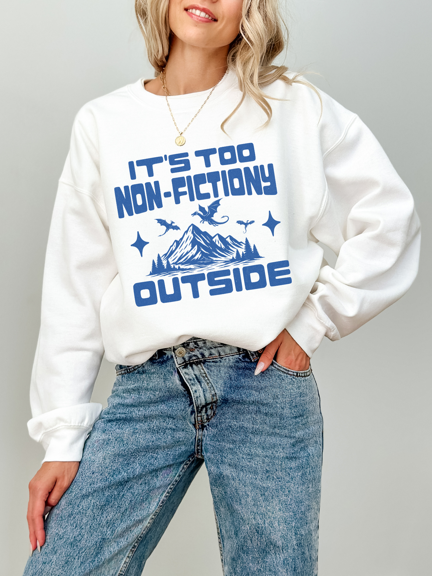 A woman wearing a white crewneck sweatshirt with a blue graphic print that reads 'IT'S TOO NON-FICTIONY OUTSIDE' with a mountain and dragon design, paired with blue jeans against a light gray background.