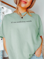 Woman wearing bay green Comfort Colors shirt that says it's too nonfictiony outside styled with gold necklace and casual outfit in bright indoor setting