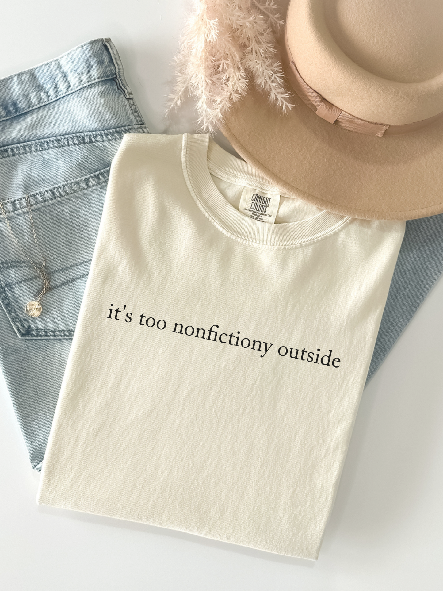 Ivory Comfort Colors book lover shirt with quote it's too nonfictiony outside styled in flatlay with jeans beige hat necklace and neutral aesthetic