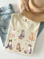 Flat lay of ivory Comfort Colors t shirt with multiple cats sitting on books and flowers cottagecore book lover design styled with jeans necklace and beige hat