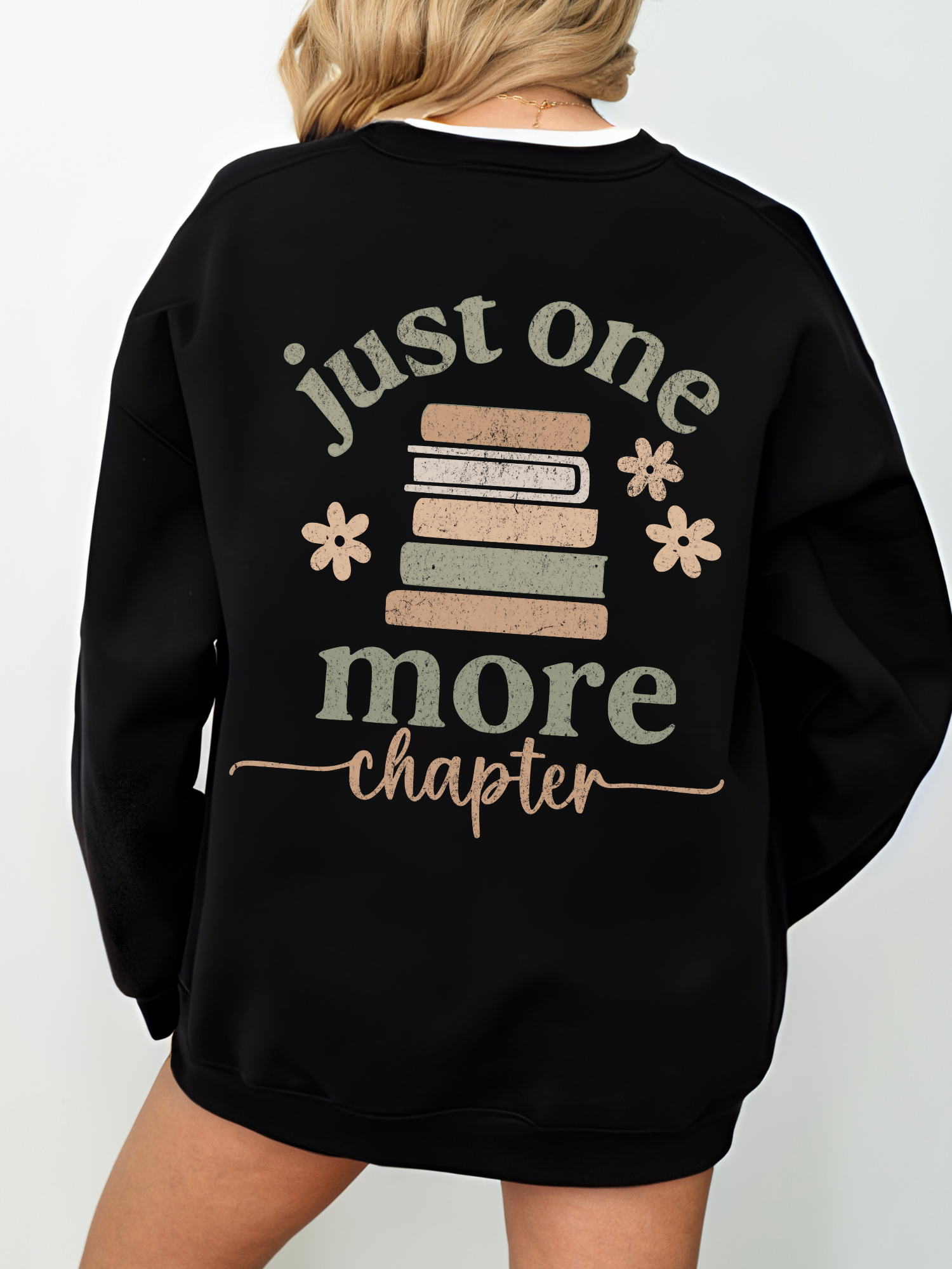 Close-up of the back of a black sweatshirt featuring a large vintage-inspired book stack design surrounded by soft florals and the phrase just one more chapter in textured cream and beige ink. Worn with layered white tee.