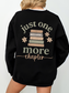 Close-up of the back of a black sweatshirt featuring a large vintage-inspired book stack design surrounded by soft florals and the phrase just one more chapter in textured cream and beige ink. Worn with layered white tee.