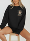 Blonde woman seated on a wooden stool wearing a black sweatshirt with a subtle front design of pastel book stack and just one more chapter quote in a cozy serif and script mix. Styled with black shorts and soft lighting.